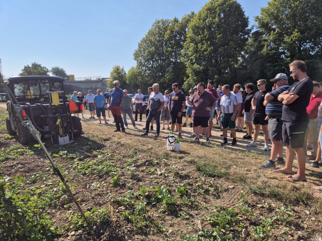 Udeleženci sestanka stojijo skupaj na polju. in opazujejo delovanje stroja.